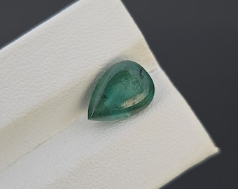 May include: A pear-shaped, polished emerald gemstone, displaying a deep green hue. The gem is set against a white surface, with a dark gray background. The gemstone is likely for jewelry making or collecting.
