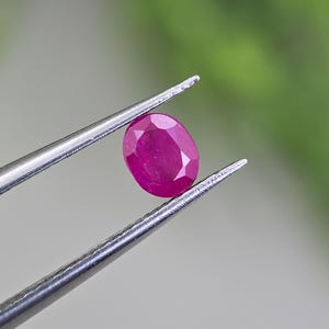 Puede incluir: Una gema de rubí de corte ovalado, con un tono rojo rosado intenso y una superficie facetada, sostenida firmemente por unas pinzas de metal plateado. La gema muestra una apariencia clara y pulida con reflejos de luz en sus facetas.