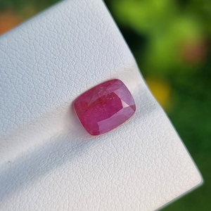 May include: A faceted, cushion-cut ruby gemstone. The ruby is a deep, saturated red colour and is displayed on a white, textured surface. The background is a blurred view of green foliage.
