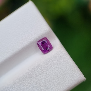 May include: A vibrant, cushion-cut, purplish-pink gemstone. The gem is set against a white, textured surface, with a blurred green background. The gemstone's facets reflect light, showcasing its clarity and color. Ideal for jewelry.