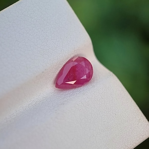 May include: A faceted, pear-shaped ruby gemstone. The ruby is a deep, vibrant red colour and is set against a white, textured surface. The gemstone is the focal point, showcasing its clarity and cut.