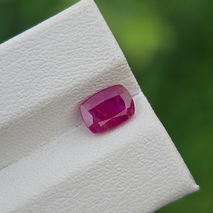 Pode incluir: Uma vibrante gema de rubi com corte almofada, exibindo um tom vermelho profundo. A gema é apresentada contra uma superfície branca texturizada, com um fundo verde desfocado. As facetas do rubi capturam a luz, mostrando seu brilho.