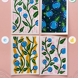 May include: Four rectangular art prints featuring blueberry illustrations. Print A has a pink dashed background. Print B has a dark blue background with white stars. Print C has a yellow wavy background. Print D has a pink checkered background.