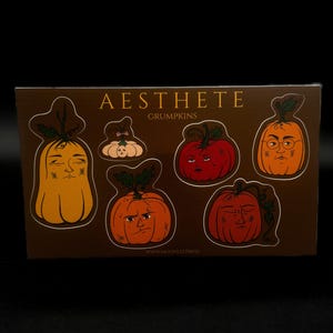 May include: A brown card featuring several pumpkin-shaped stickers with various facial expressions. The stickers are in shades of orange, red, and yellow, with green leaves. The text "Aesthete Grumpkins" is printed at the top, and "www.moonlitpress.com" at the bottom.