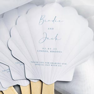May include: A set of white shell-shaped paper fans with wooden handles. The text reads: Brodie AND Jack, 05.05.25 LINDOS, RHODES, THANK YOU FOR SHARING OUR SPECIAL DAY WITH US! The design features light blue script and block lettering on a scalloped surface.