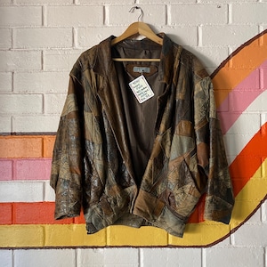 May include: A brown leather jacket featuring a patchwork design of various leather shades and textures. The jacket has a collared neckline, long sleeves, and a front zip closure. A white tag with handwritten text is attached to the inner collar.