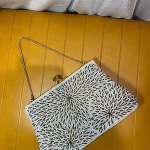 May include: A rectangular handbag featuring a white and gold beaded pattern arranged in circular floral-like motifs. The bag includes a gold-toned metal frame, a top clasp closure, and a thin gold-toned metal chain strap for carrying.