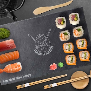 May include: A black slate serving board with a variety of sushi rolls, including salmon, tuna, and shrimp. The board also features a logo for "Sushi Bar John & Carol" and the text "You Maki Miso Happy".