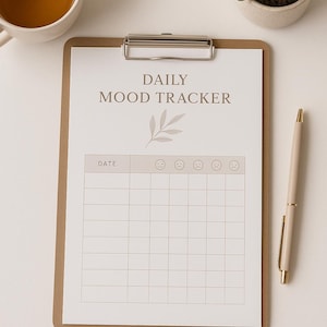 Puede incluir: Un portapapeles con una hoja de "Daily Mood Tracker", un bolígrafo, una taza de té y una pequeña planta en maceta. El rastreador de estado de ánimo tiene espacios para fechas y emoticonos. El bolígrafo es beige con detalles dorados.