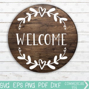 May include: A round wooden sign with the word "WELCOME" in white lettering. The sign is decorated with a white wreath of leaves and hearts.