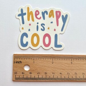 May include: A sticker with the text "therapy is cool" in blue, yellow, and pink lettering with small pink stars. The sticker is about 2 inches wide.