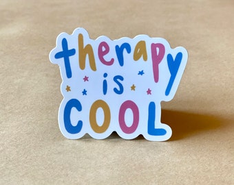 May include: A white sticker with the text "therapy is cool" in colorful, stylized lettering. The text is in blue, pink, and gold. There are small white stars scattered around the text.