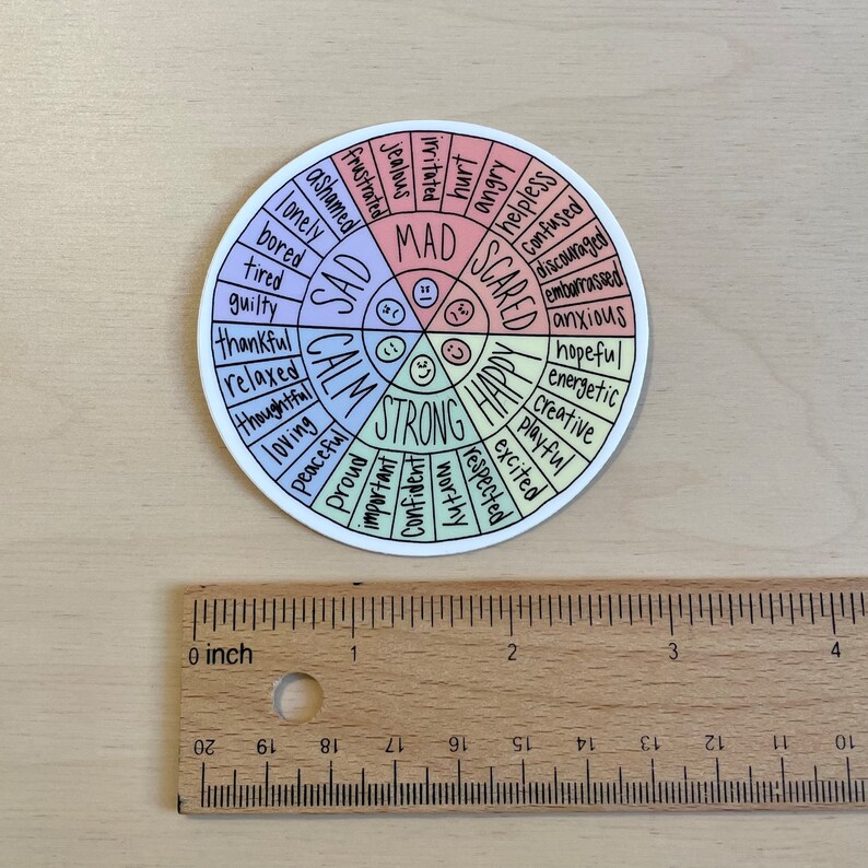 May include: A white circular sticker with a colourful wheel of emotions. The wheel is divided into four sections:  "Sad", "Calm", "Strong", and "Happy". Each section has a list of words describing different emotions. The sticker is 5 cm in diameter.