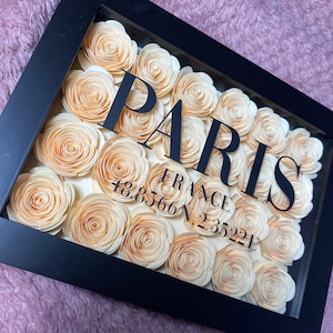 May include: A black framed shadow box with the word "PARIS" in black lettering and the coordinates "48.8566 N, 2.3522 E" below. The box is filled with peach-coloured paper flowers.