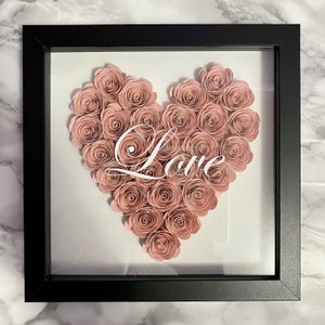 May include: A black framed shadow box with a pink paper flower heart inside. The heart is made of many small paper roses and the word "Love" is written in white inside the heart.