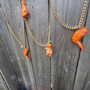 May include: Gold-toned chain necklaces with orange chicken wing pendants hang against a weathered wooden fence. The necklaces feature various wing shapes, creating a unique and playful accessory. The background includes a glimpse of colorful clothing.