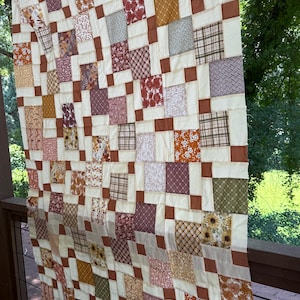 May include: A patchwork quilt featuring a grid of alternating cream-colored squares and various patterned fabric squares in shades of rust, brown, olive green, and gold. The quilt displays a variety of floral, plaid, and geometric prints throughout.