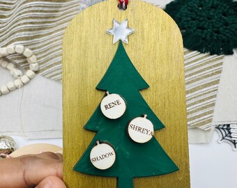 Könnte beinhalten: Weihnachtsbaumschmuck aus Holz mit goldenem Hintergrund und grünem Baum. Der Baum hat drei runde Kugeln mit den Namen "Rene", "Shreya" und "Shadow". Ein silberner Stern ziert die Spitze.