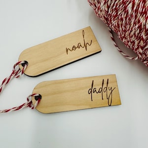 May include: Two wooden gift tags, one with "noah" and the other with "daddy" engraved in cursive. Each tag has a hole for string and is attached to red and white twine. A spool of red, white, and gold twine is in the background.