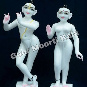 May include: White marble statue of a couple standing on separate bases. The male figure is on the left and the female figure is on the right. The figures are both nude and have painted details on their faces. The male figure has a yellow line on his chest and the female figure has a red dot on her forehead. The text "Gour Moorti Kala" is visible in the image.