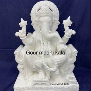 Op de afbeelding: Wit marmeren beeld van Ganesha, de hindoe&iuml;stische god van wijsheid en voorspoed, zittend op een troon. Het beeld is ingewikkeld gebeeldhouwd met details en heeft een traditioneel ontwerp. Het beeld is op de basis ge&euml;tiketteerd met "Gour moorti kala".