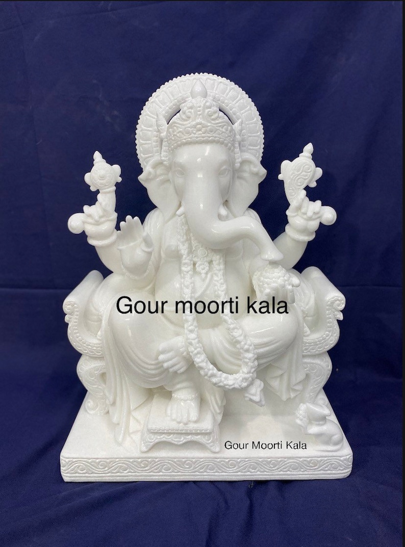 Op de afbeelding: Wit marmeren beeld van Ganesha, de hindoe&iuml;stische god van wijsheid en voorspoed, zittend op een troon. Het beeld is ingewikkeld gebeeldhouwd met details en heeft een traditioneel ontwerp. Het beeld is op de basis ge&euml;tiketteerd met "Gour moorti kala".