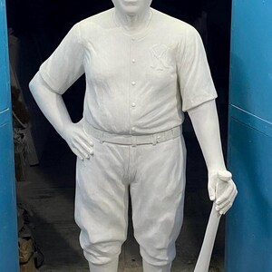 May include: A life-sized white statue of a baseball player wearing a button-down jersey with a NY logo on the chest, knicker-style pants, and cleats. The figure stands on a rectangular base, holding a baseball bat in one hand with the other hand on a hip.