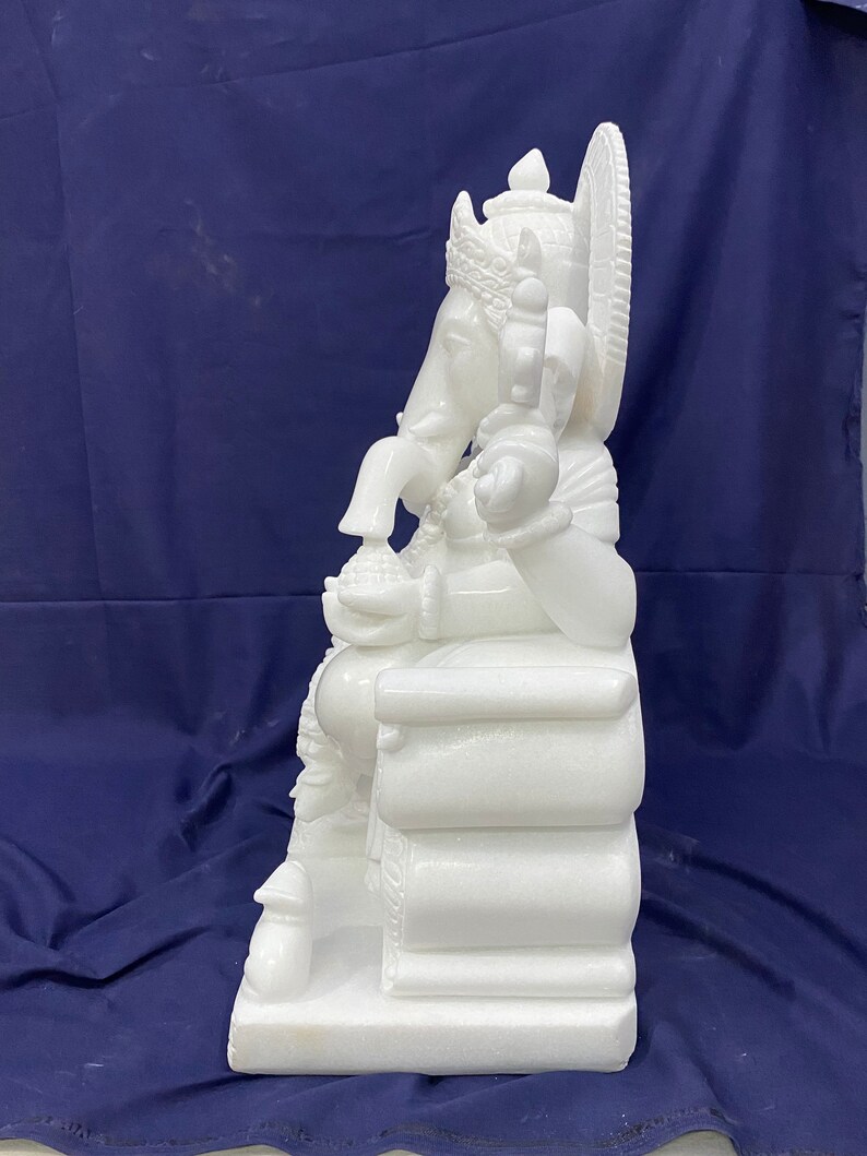 Op de afbeelding: Wit marmeren beeld van Ganesha, de hindoe&iuml;stische god van wijsheid en voorspoed, zittend op een troon. Het beeld is ingewikkeld gedetailleerd en heeft een kroon, een slurf en een grote buik.