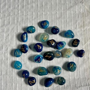 May include: A collection of blue and green agate stones with gold runes painted on them. The stones are arranged in a circle on a white fabric background.