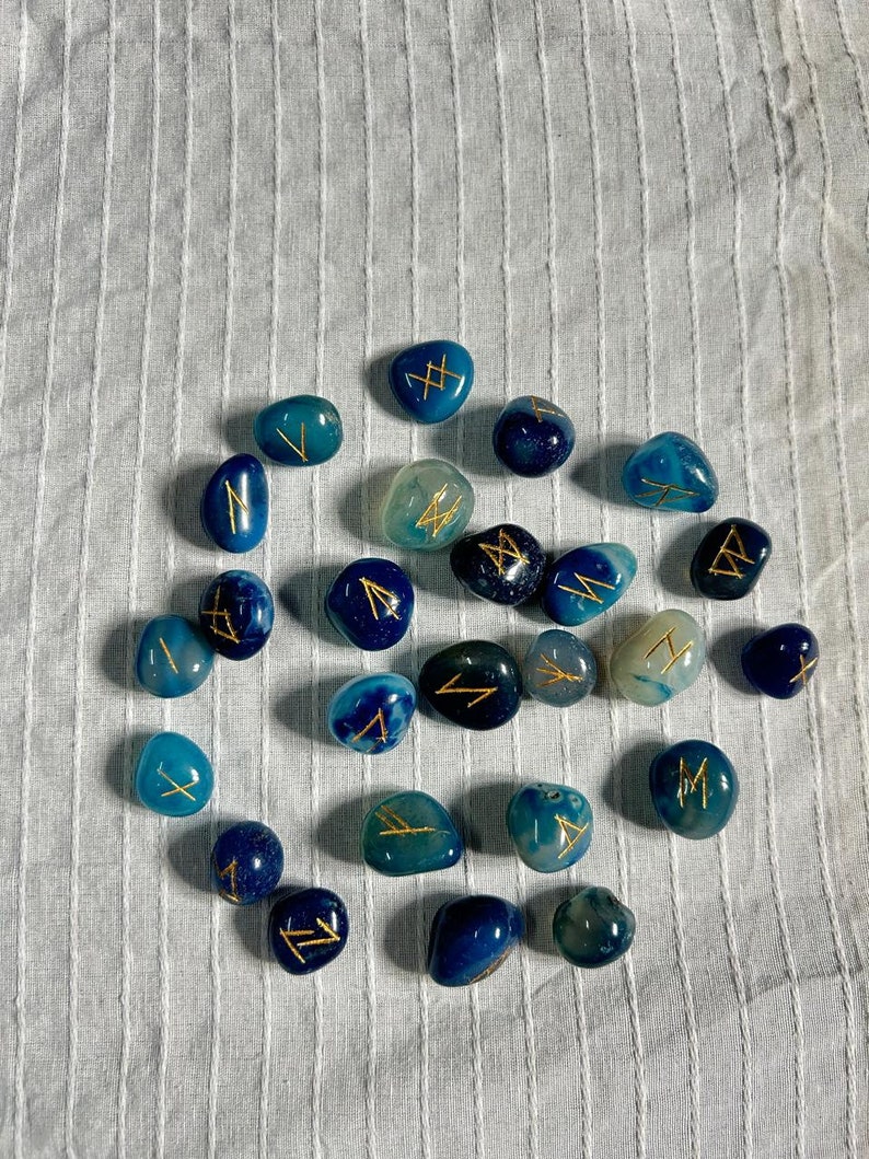 May include: A collection of blue and green agate stones with gold runes painted on them. The stones are arranged in a circle on a white fabric background.