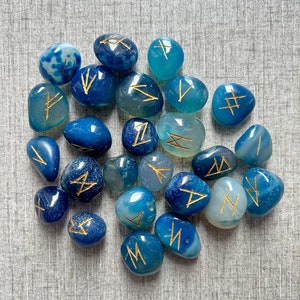 May include: A collection of blue agate stones with gold runes painted on them. The stones are tumbled and smooth.