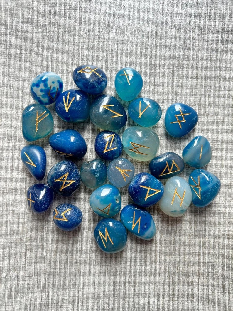 May include: A collection of blue agate stones with gold runes painted on them. The stones are tumbled and smooth.