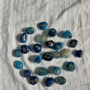 May include: A collection of blue agate stones with gold runes inscribed on them. The stones are arranged in a circle on a white fabric background.