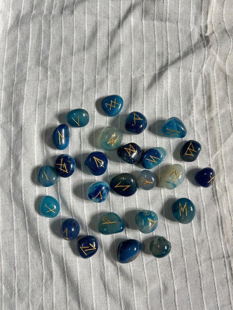 May include: A collection of blue agate stones with gold runes inscribed on them. The stones are arranged in a circle on a white fabric background.