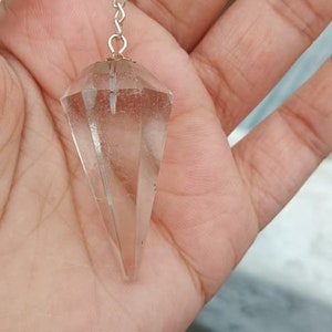 Könnte beinhalten: Ein klares Quarzkristall-Pendel mit einer silbernen Kette. Das Pendel hat die Form eines spitzen Tropfens.