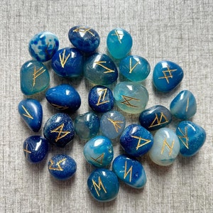 May include: A collection of blue and white agate stones with gold runes inscribed on them. The stones are tumbled and smooth.