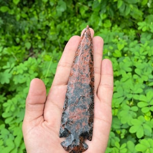 Puede incluir: Una punta de flecha de color marrón rojizo con motas negras, sostenida en la mano. La punta de flecha es de piedra y tiene una punta afilada y una base con muescas. El fondo es una vista borrosa de follaje verde.
