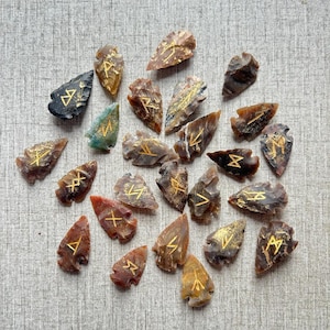 May include: A collection of 20 arrowhead-shaped stones with gold runes carved into them. The stones are various shades of brown, black, and green.