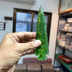 May include: A green glass arrowhead with a textured surface. The arrowhead is pointed at the top and has a rounded base.
