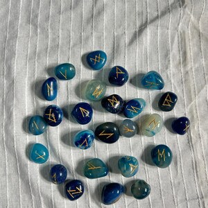 May include: A set of 20 blue agate stones with gold runes painted on them. The stones are scattered on a white fabric background.