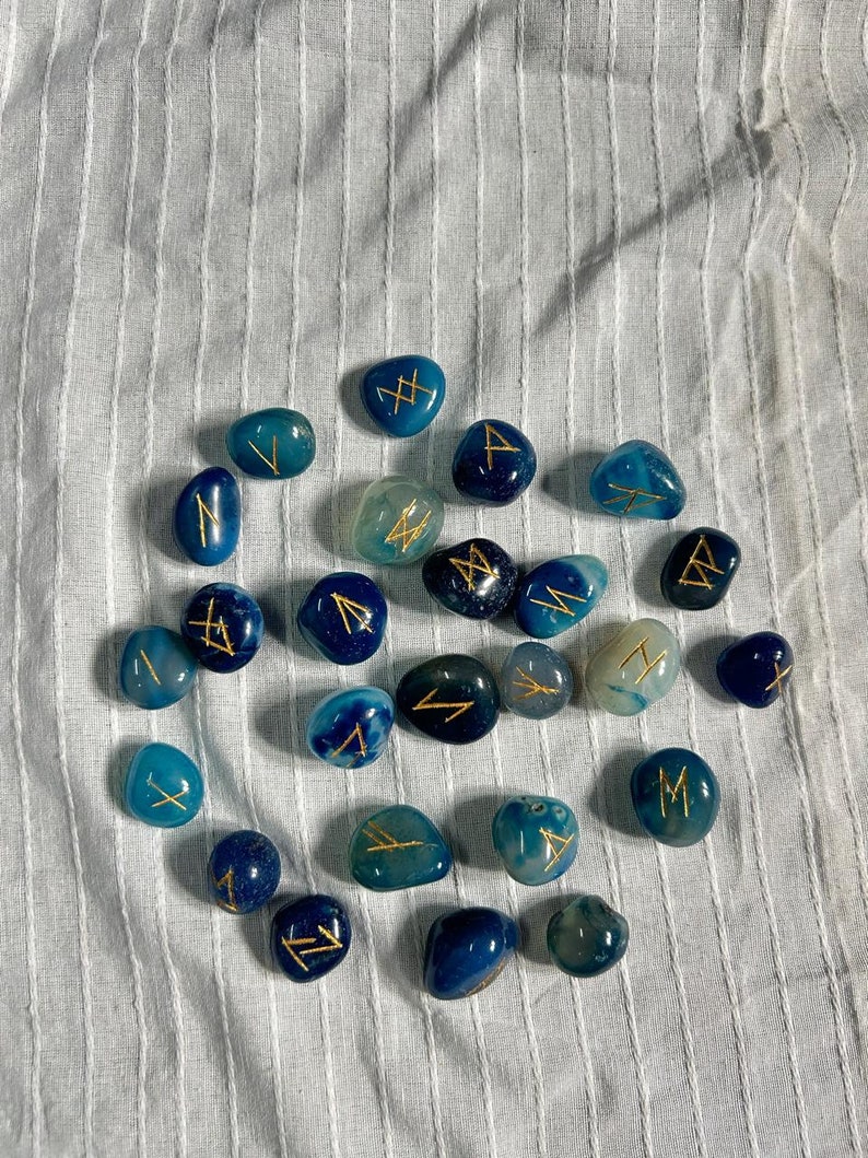 May include: A set of 20 blue agate stones with gold runes painted on them. The stones are scattered on a white fabric background.
