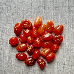 May include: A collection of 20 carnelian stones with gold runes inscribed on each. The stones are arranged in a loose pile on a grey textured surface.
