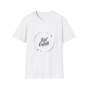 May include: White t-shirt with a crew neck and short sleeves. The shirt features a black circular design with the words "Lead by Example" in a stylized script. Small star shapes are around the circle.