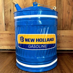 May include: A blue metal gasoline can with white stripes and a wooden handle. The can features a yellow and blue label with the words "NEW HOLLAND GASOLINE". The can has a spout and a lid with a metal handle.