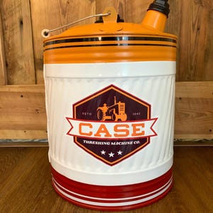 May include: A vintage-style metal container with an orange top and a white body. The container features black stripes, a red base, and a logo that reads "CASE Threshing Machine Co." with a tractor illustration. A metal handle is attached.