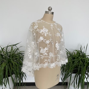 May include: White lace bridal bolero with a floral pattern. The bolero has a round neckline and 3/4 sleeves. The lace is delicate and has a scalloped edge.