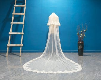 May include: A long, ivory-colored bridal veil with delicate lace trim. The veil is displayed on a mannequin against a vibrant blue backdrop. A wooden ladder and a decorative vase with flowers are also visible.