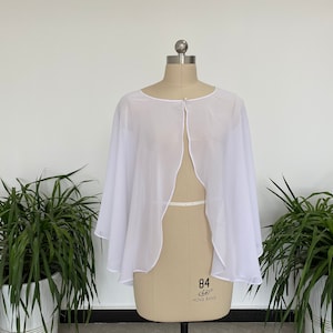 Op de afbeelding: Witte, doorschijnende cape met een ronde halslijn en een ritssluiting in het midden van de rug. De cape wordt gepresenteerd op een mannequin.