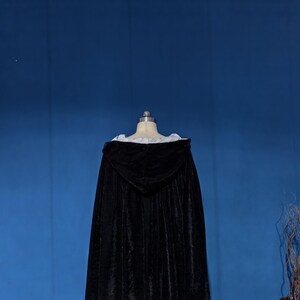 May include: A floor-length black velvet cloak featuring a hood and a white interior lining. The garment is displayed on a mannequin, showcasing the dark fabric texture, the structured hood, and the contrasting light-colored trim along the neckline.