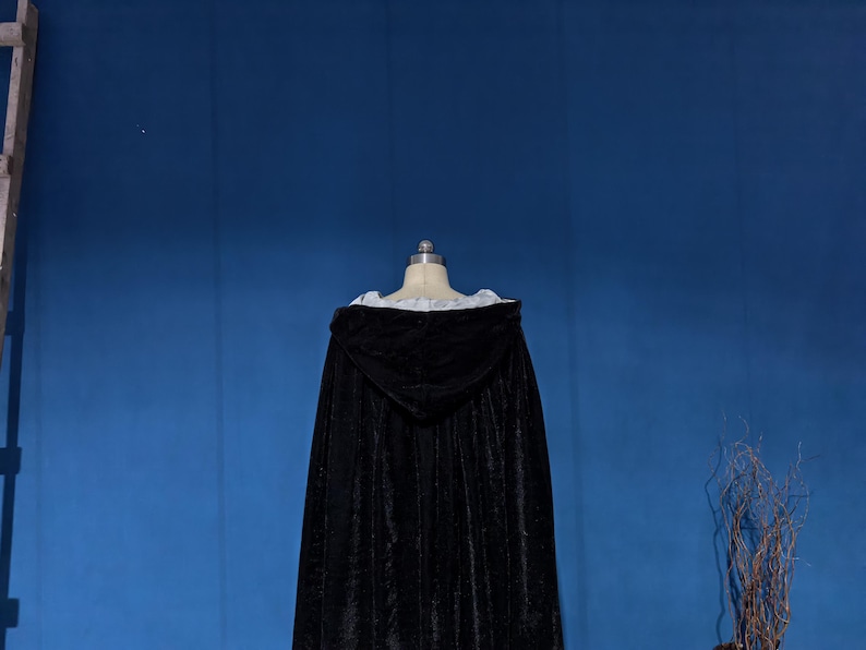 May include: A floor-length black velvet cloak featuring a hood and a white interior lining. The garment is displayed on a mannequin, showcasing the dark fabric texture, the structured hood, and the contrasting light-colored trim along the neckline.
